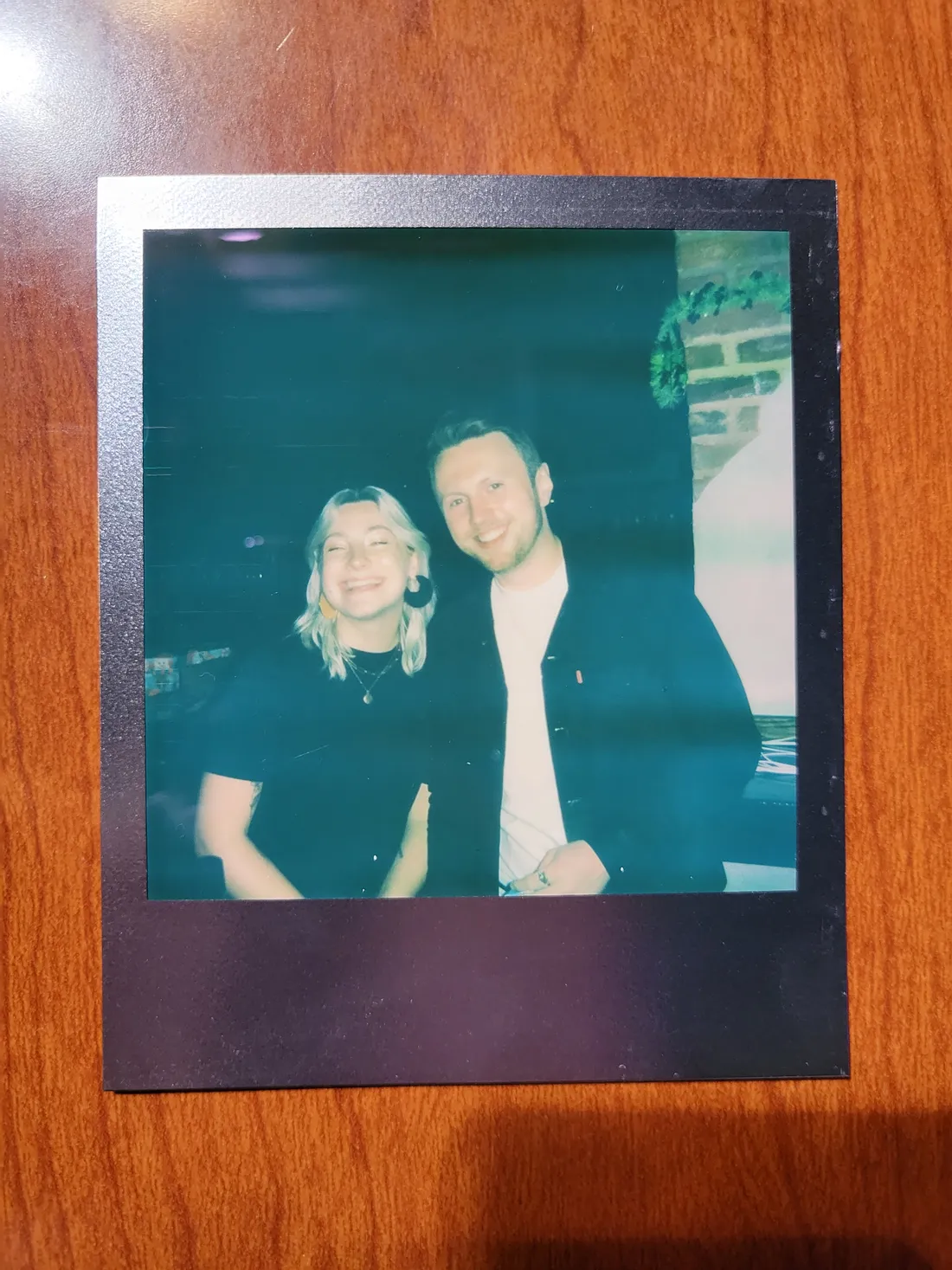 A Polaroid on a wooden table: Cat and Zach smiling side by side at a party.