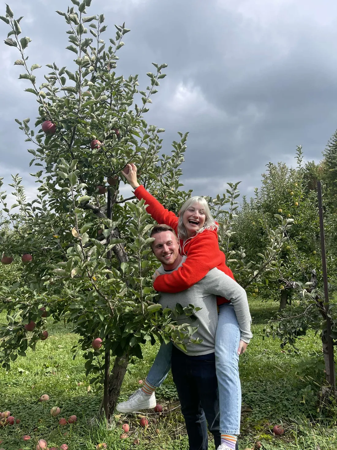 Cat riding on Zach’s back in an apple orchard, laughing as she reaches for an apple.